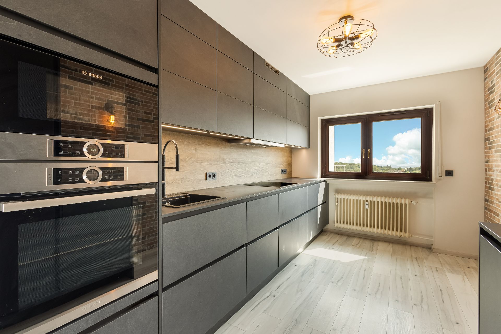 Moderne Küche mit grauen Schränken, Einbaugeräten, großem Fenster und hellem Boden.