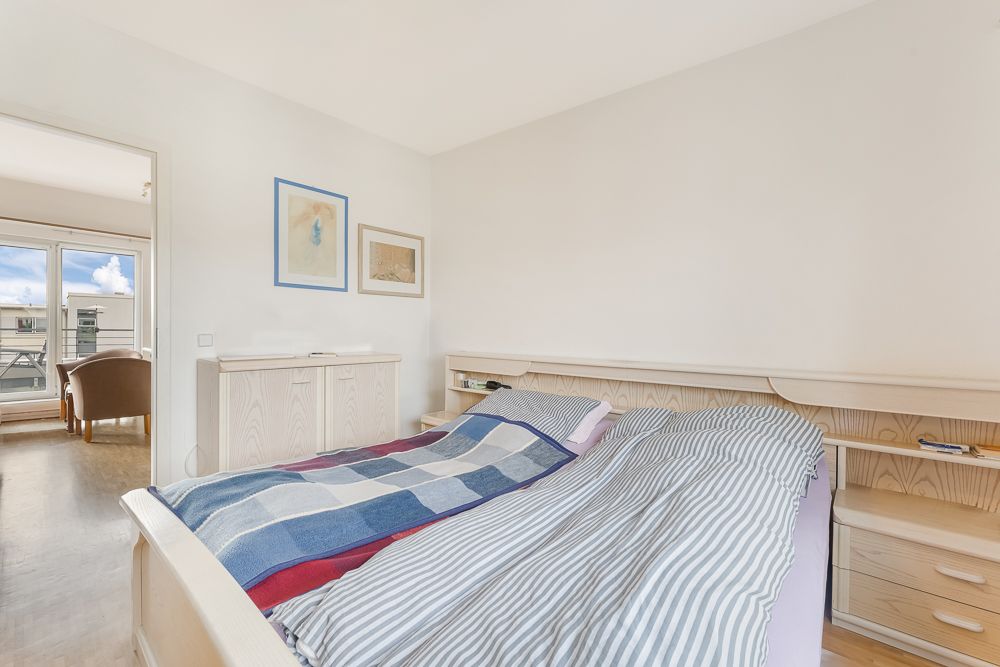 Helles Schlafzimmer mit Doppelbett, Kommode und Zugang zu Balkon durch große Glastür.