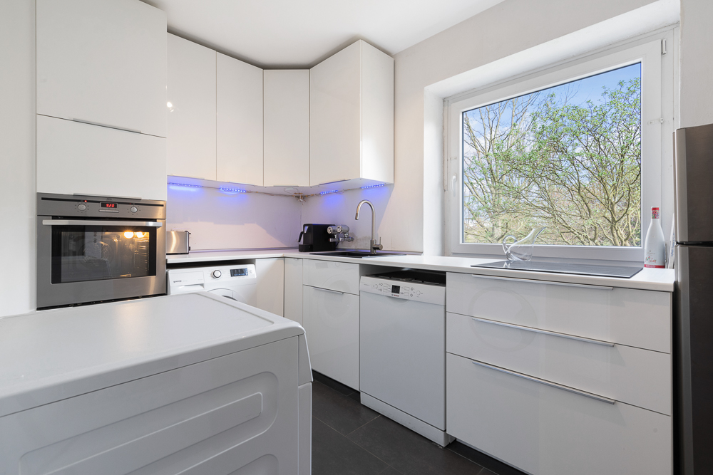Moderne weiße Küche mit großen Fenstern, Spülmaschine, Backofen und Waschmaschine.