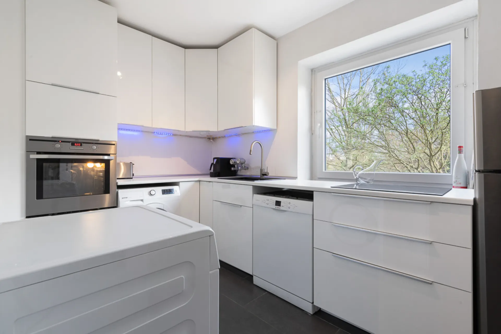 Moderne weiße Küche mit großen Fenstern, Spülmaschine, Backofen und Waschmaschine.