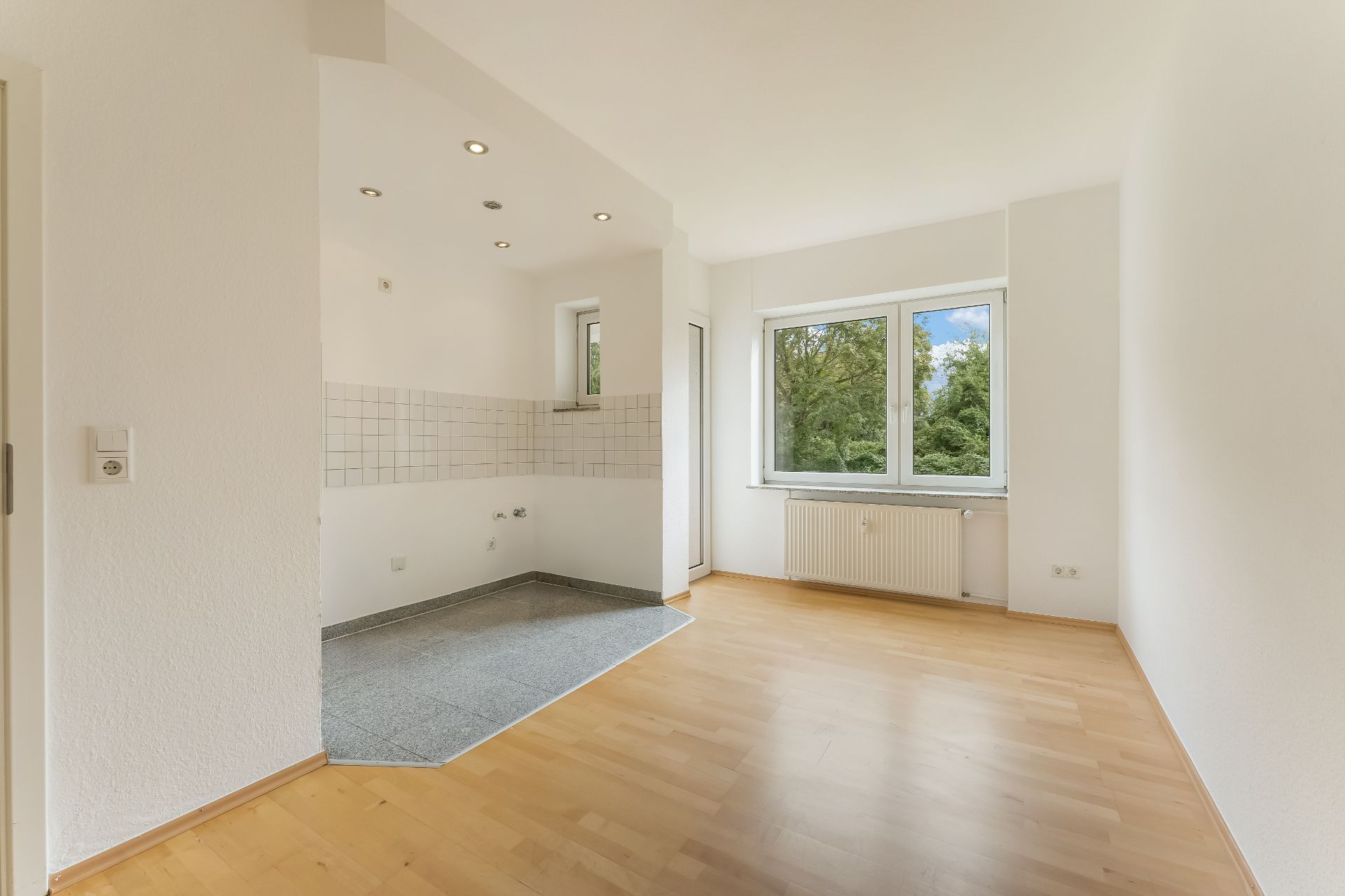 Helles, leeres Zimmer mit Parkettboden, großem Fenster und offenem Küchenbereich.