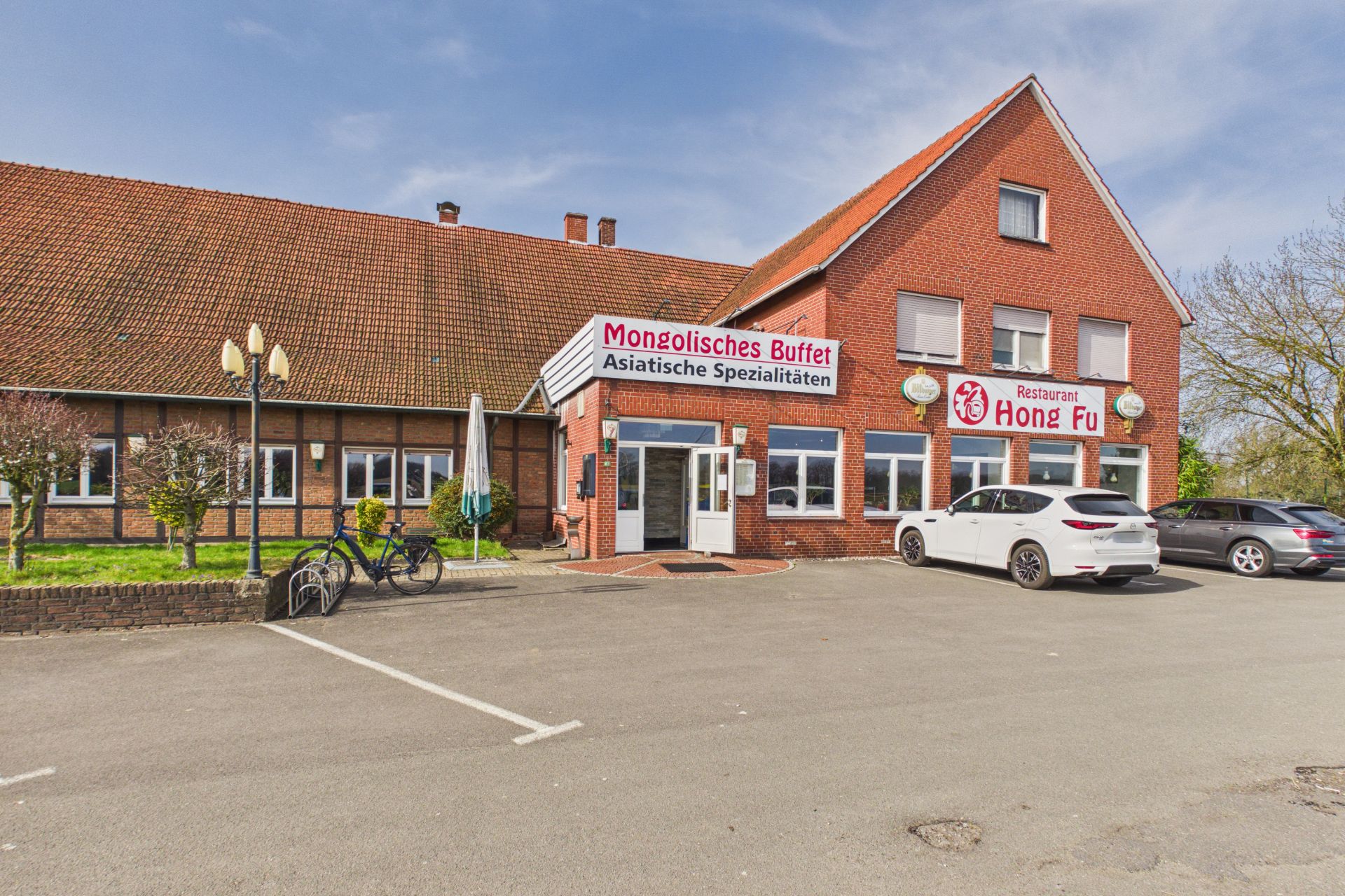 Backsteingebäude mit Restaurant, davor Parkplatz mit Autos und Fahrrädern.