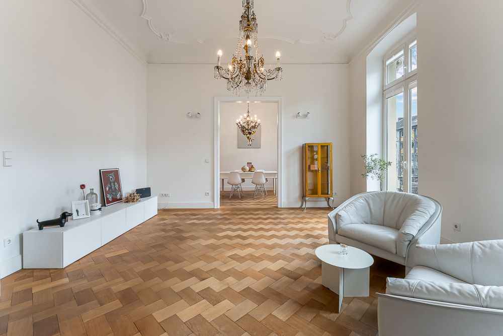 Helles Wohnzimmer mit Parkettboden, Kronleuchter, weißen Sesseln und Durchgang zum Esszimmer.