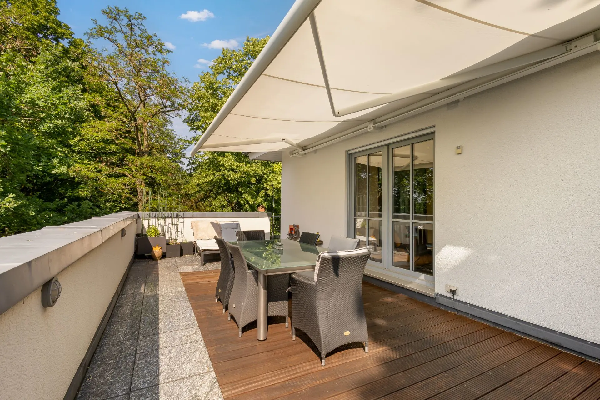 Große Terrasse mit Markise, Esstisch und Stühlen, umgeben von Bäumen.