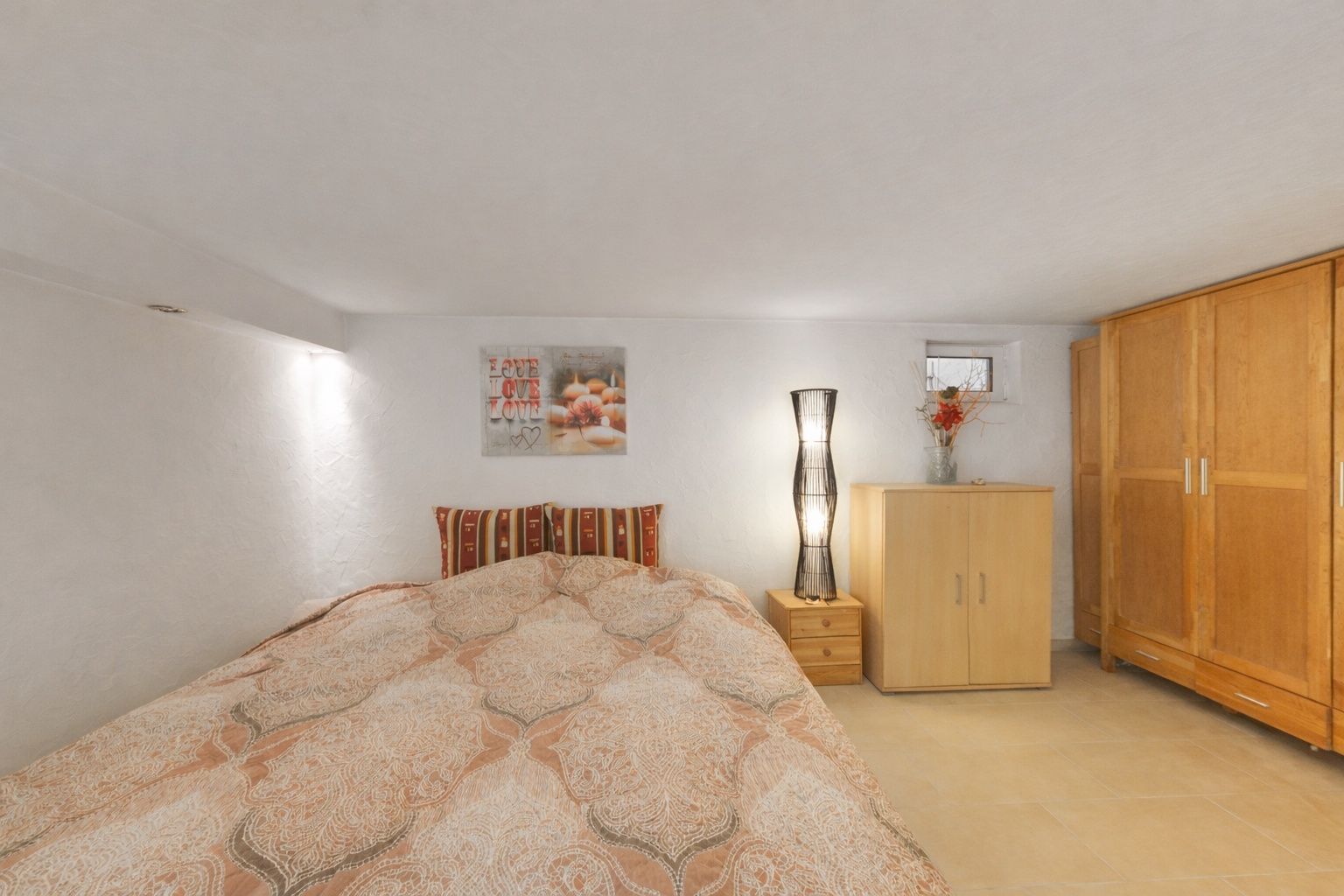 Schlafzimmer mit Doppelbett, Nachttisch, Stehlampe, Kommode, Schrank und Wanddeko.