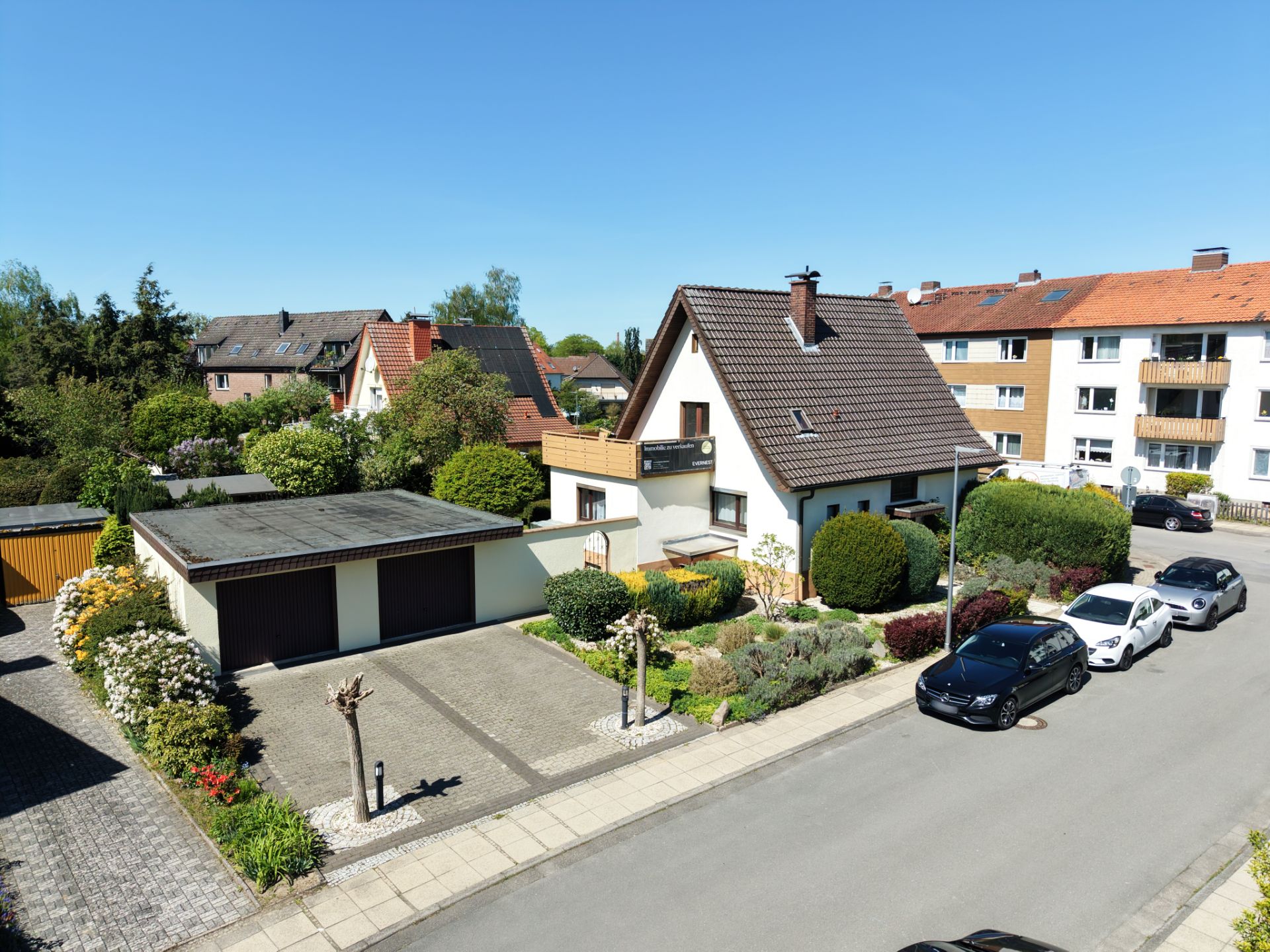 Einfamilienhaus mit Garten, Garage und geparkten Autos an der Straße bei sonnigem Wetter.