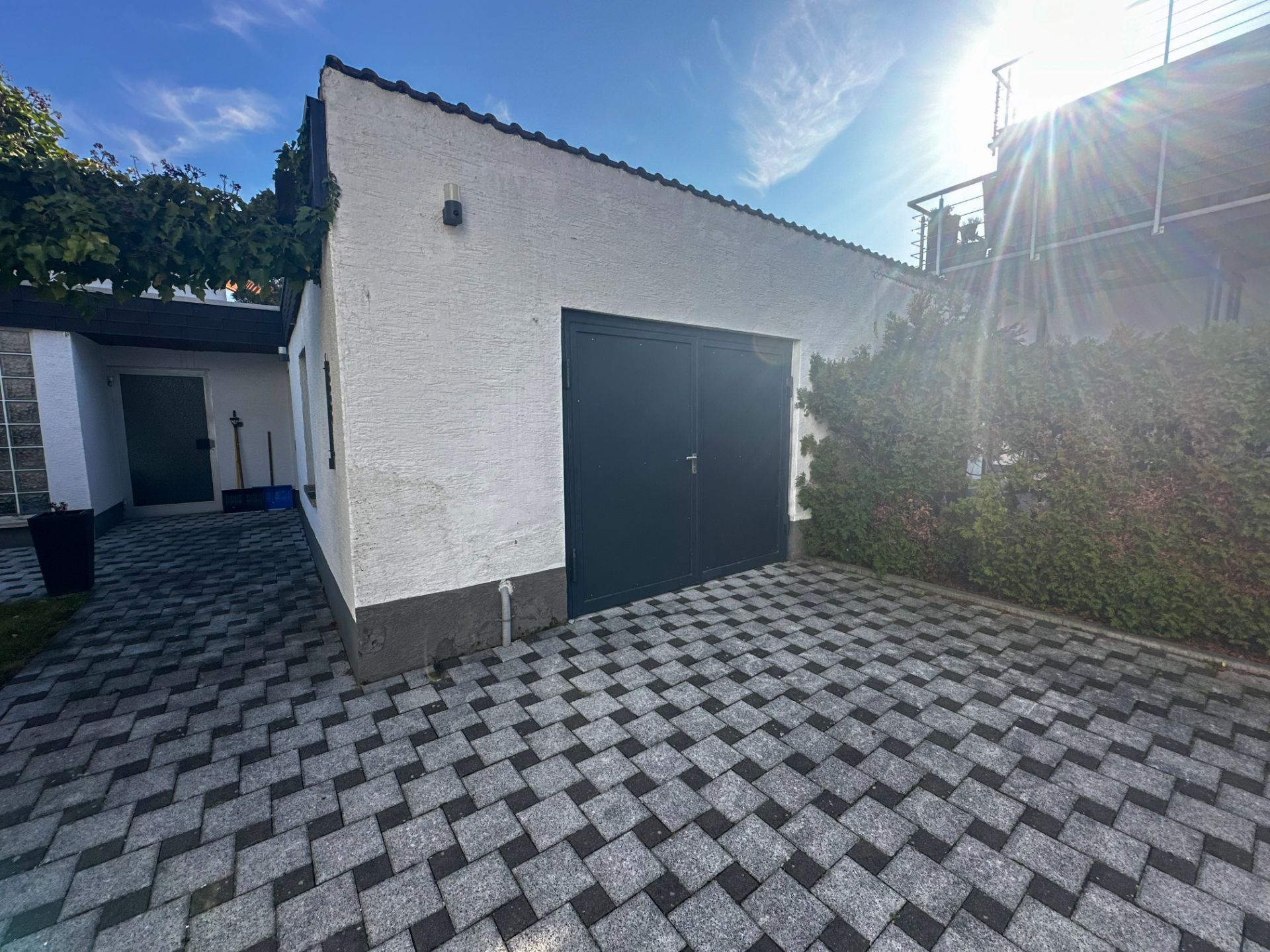 Weiße Garage mit dunklem Tor auf gepflastertem Hof, daneben grüne Hecke und Nebenhaus.