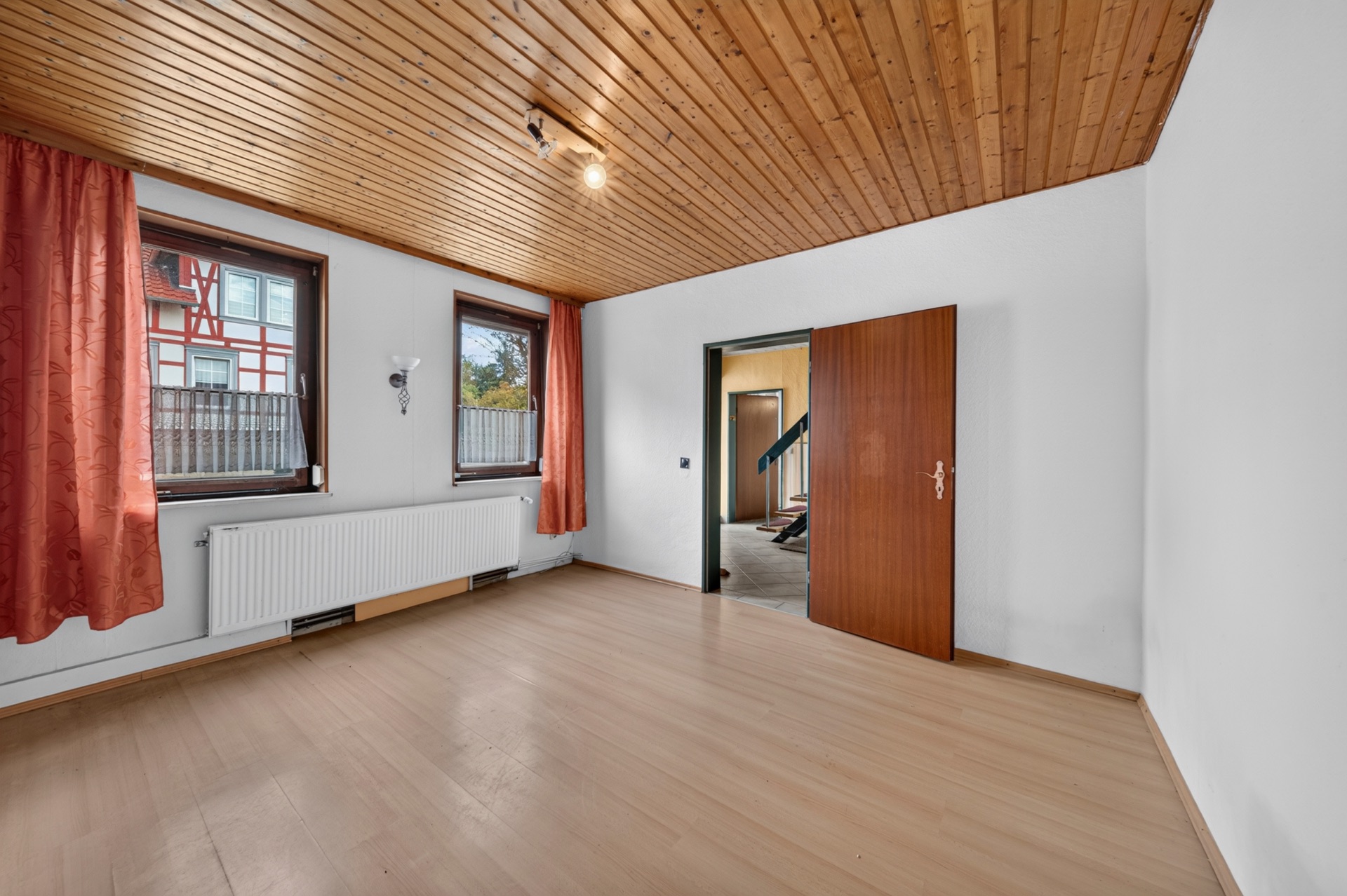 Leerer Raum mit Holzboden, Holzdecke, zwei Fenstern mit roten Vorhängen und offener Tür.