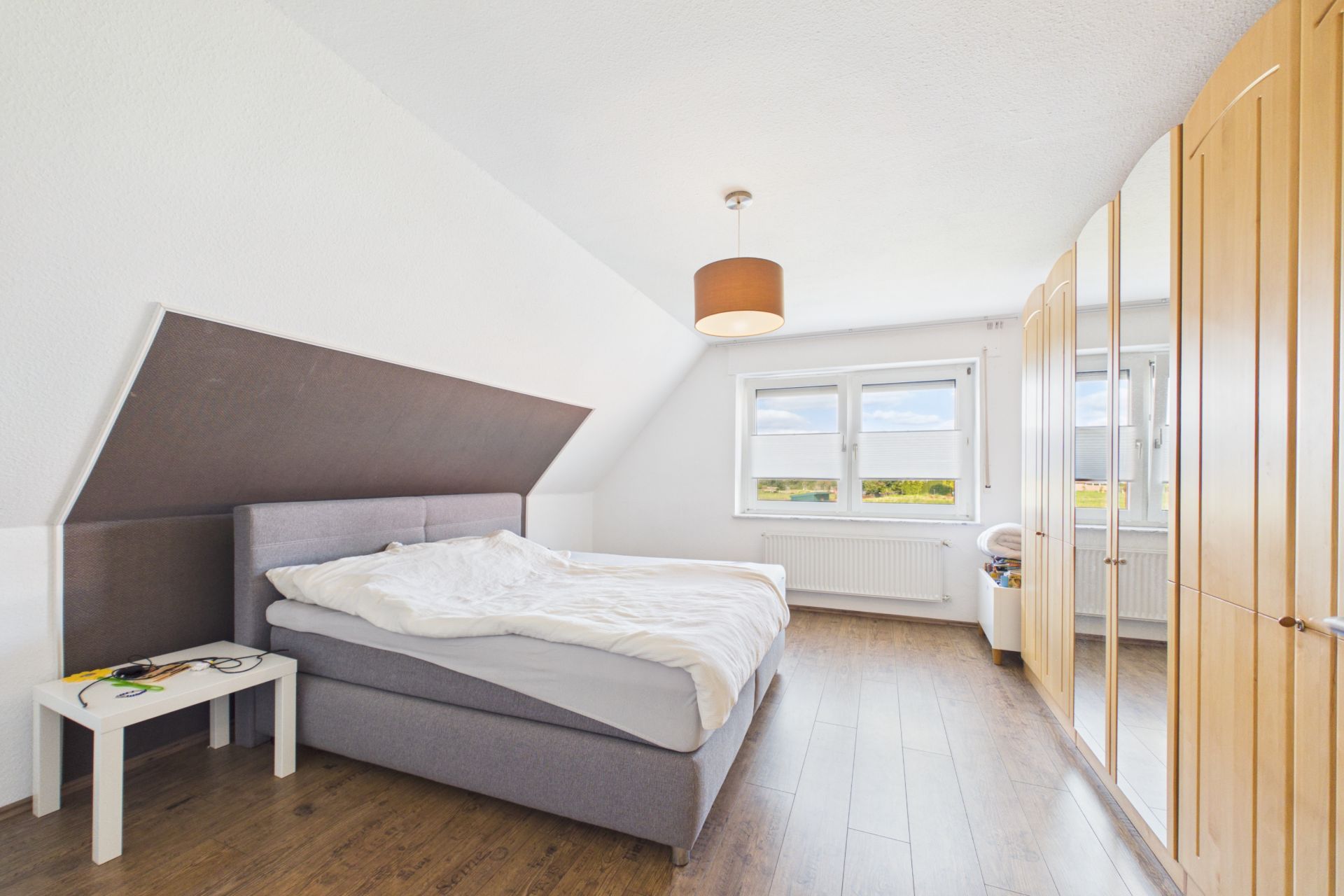 Helles Schlafzimmer mit Doppelbett, Nachttisch und großem Kleiderschrank, Fenstern und Holzboden.