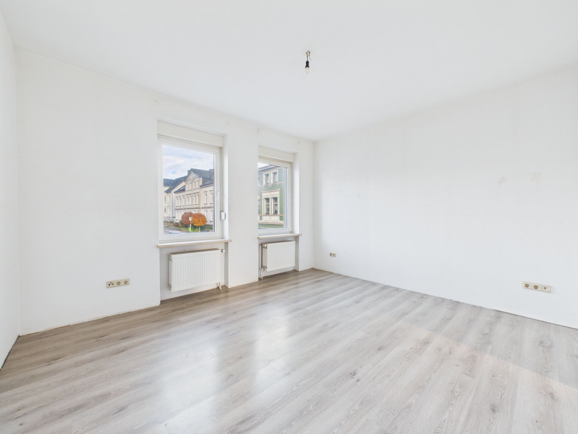 Heller, leerer Raum mit zwei Fenstern, weißen Wänden und hellem Holzfußboden.