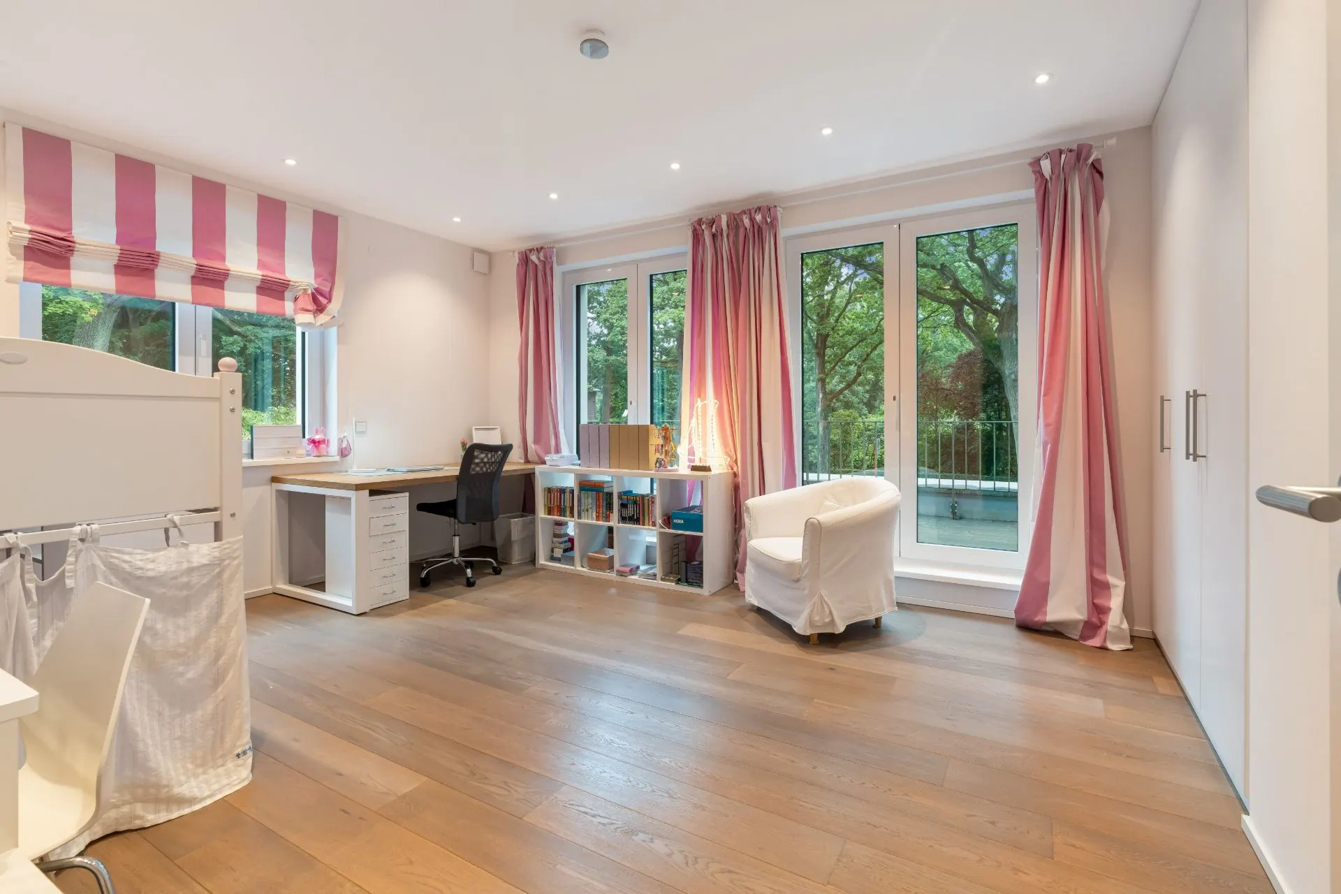 Helles Kinderzimmer mit Holzfußboden, Schreibtisch, Regal, Sessel und rosa-weißen Vorhängen.