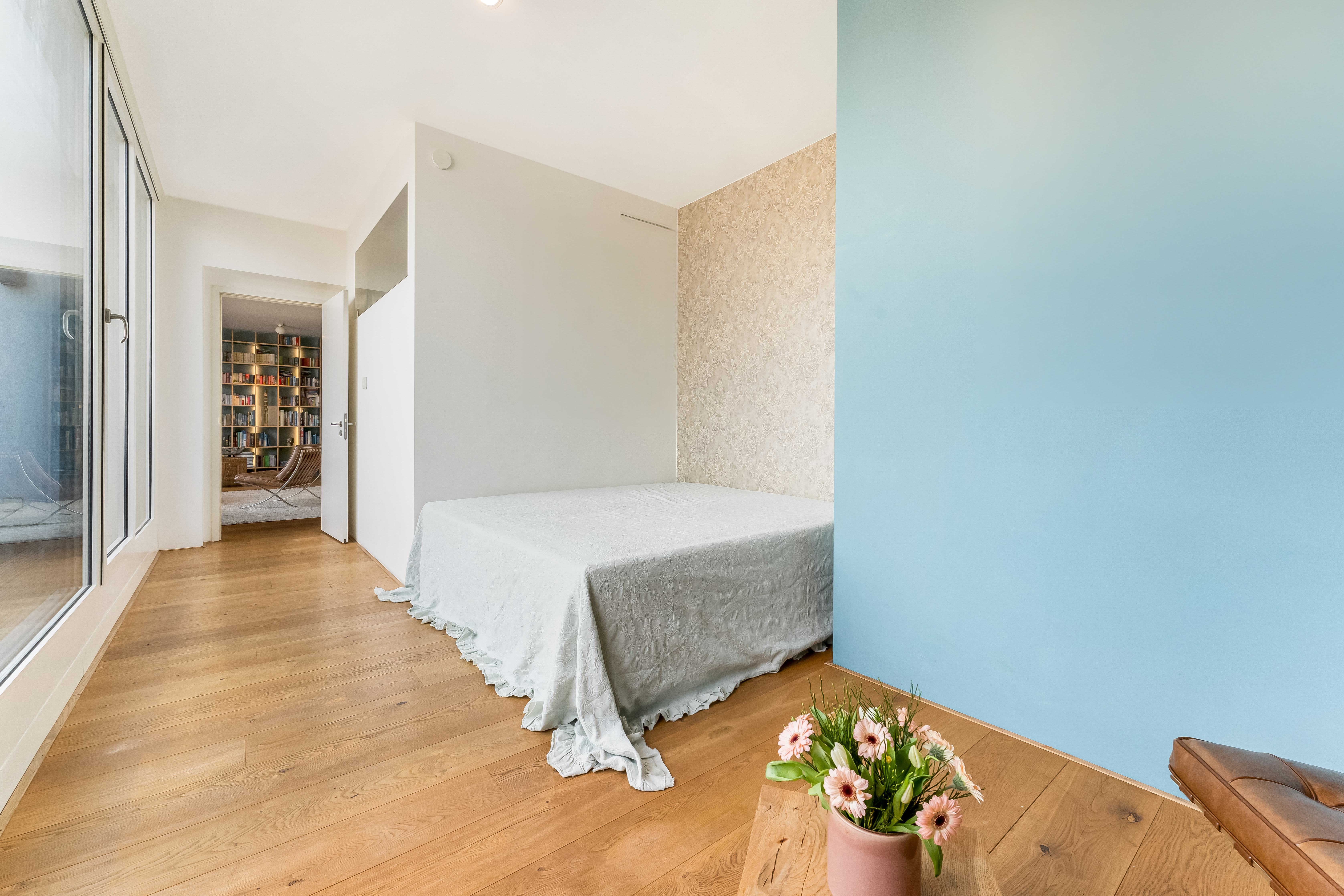 Helles Schlafzimmer mit Holzboden, Bett mit hellem Bezug und Blumen in einem Topf im Vordergrund.