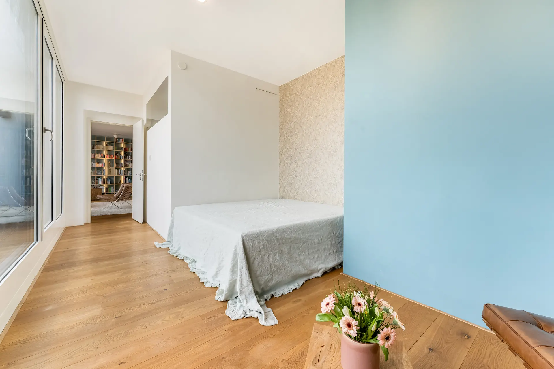 Helles Schlafzimmer mit Holzboden, Bett mit hellem Bezug und Blumen in einem Topf im Vordergrund.