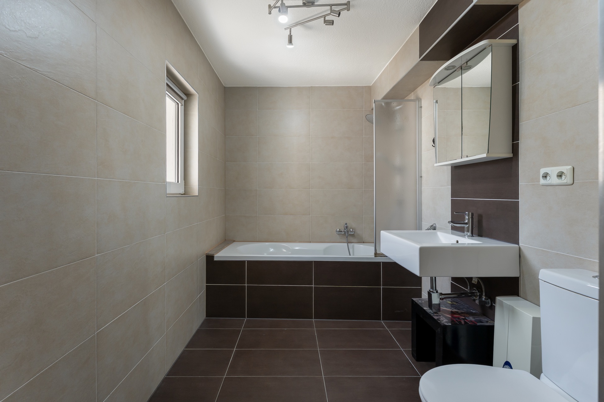 Modernes Badezimmer mit Badewanne, Waschbecken, Spiegel und WC, beige und braune Fliesen.