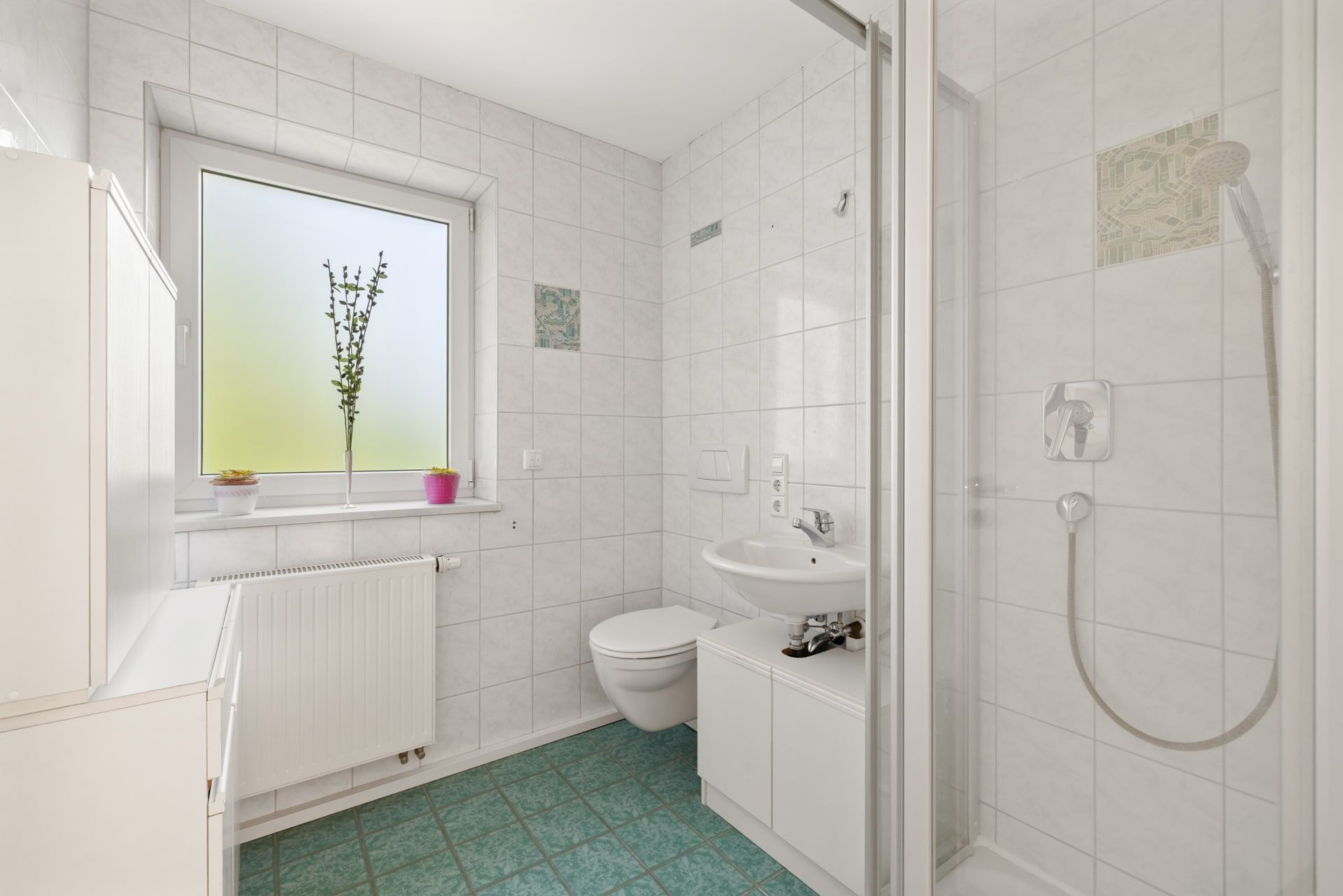 Modernes Badezimmer mit Dusche, WC, Waschbecken und Fenster mit zwei Blumentöpfen auf der Fensterbank.