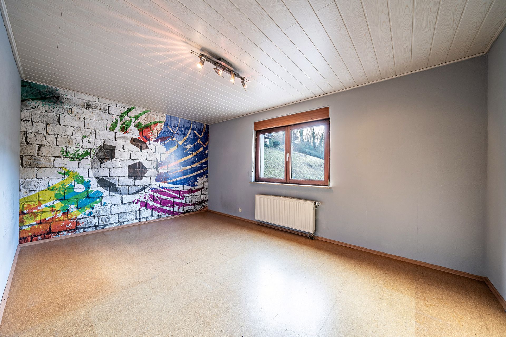 Leeres Zimmer mit großem Fenster und bunter Fußball-Grafik auf einer Wand.