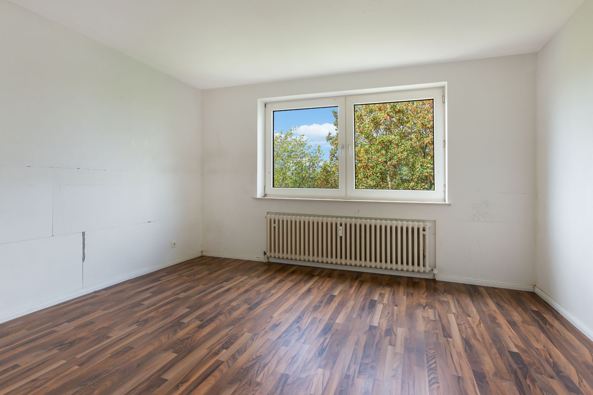 Leeres Zimmer mit Holzfußboden, weißer Wand und großem Fenster mit Blick ins Grüne.