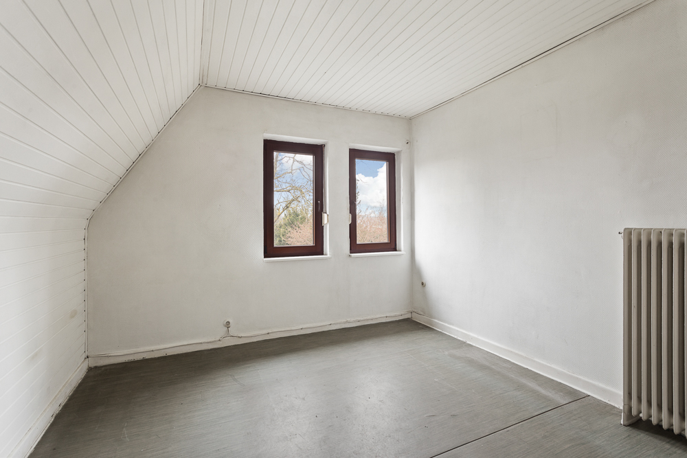 Leeres Zimmer mit weißen Wänden, zwei Fenstern und Heizkörper auf der rechten Seite.