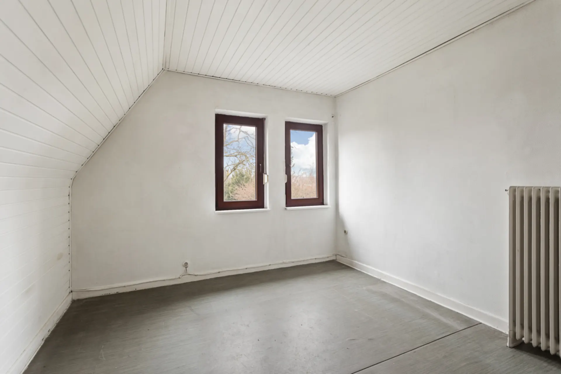 Leeres Zimmer mit weißen Wänden, zwei Fenstern und Heizkörper auf der rechten Seite.