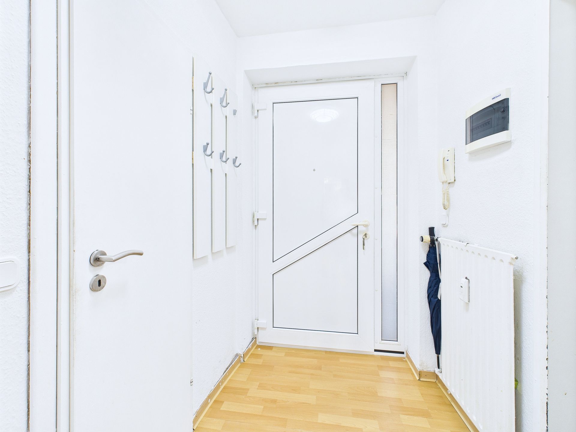 Heller Flur mit weißer Haustür, Garderobenhaken, Heizung und Holzfußboden.