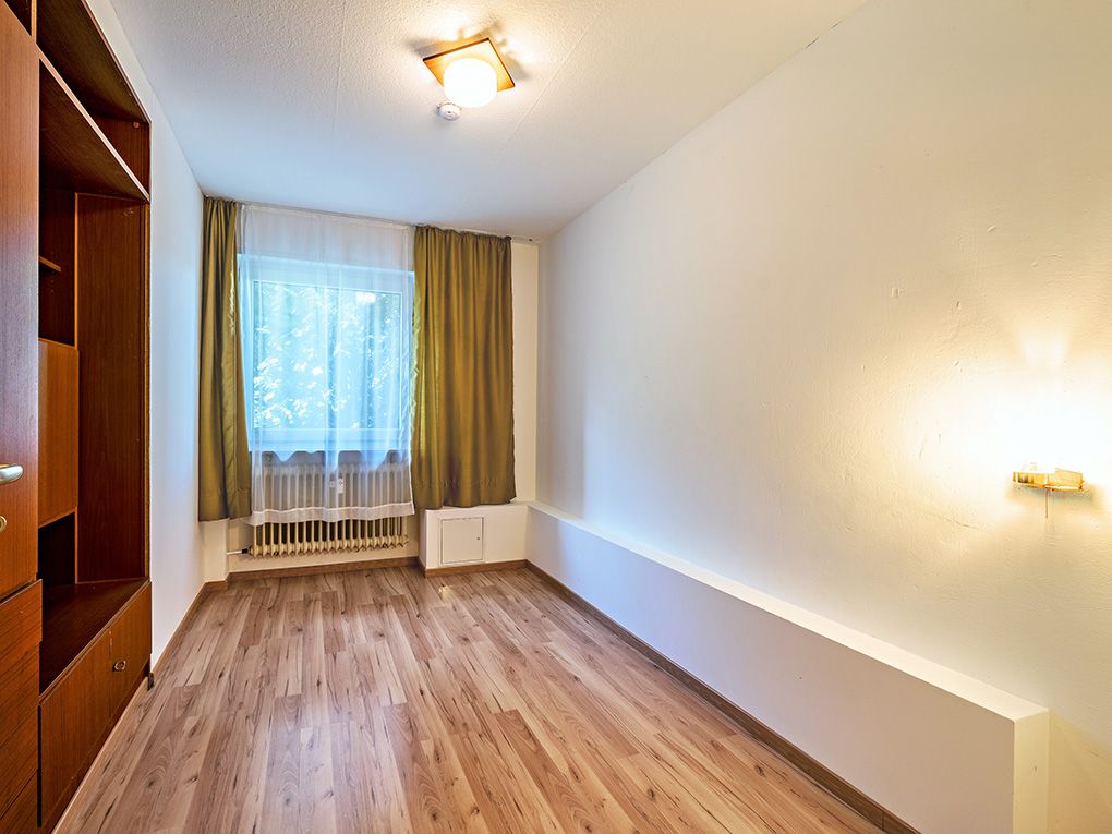 Helles, leeres Zimmer mit Holzfußboden, Fenster mit Vorhängen und Wandregal.
