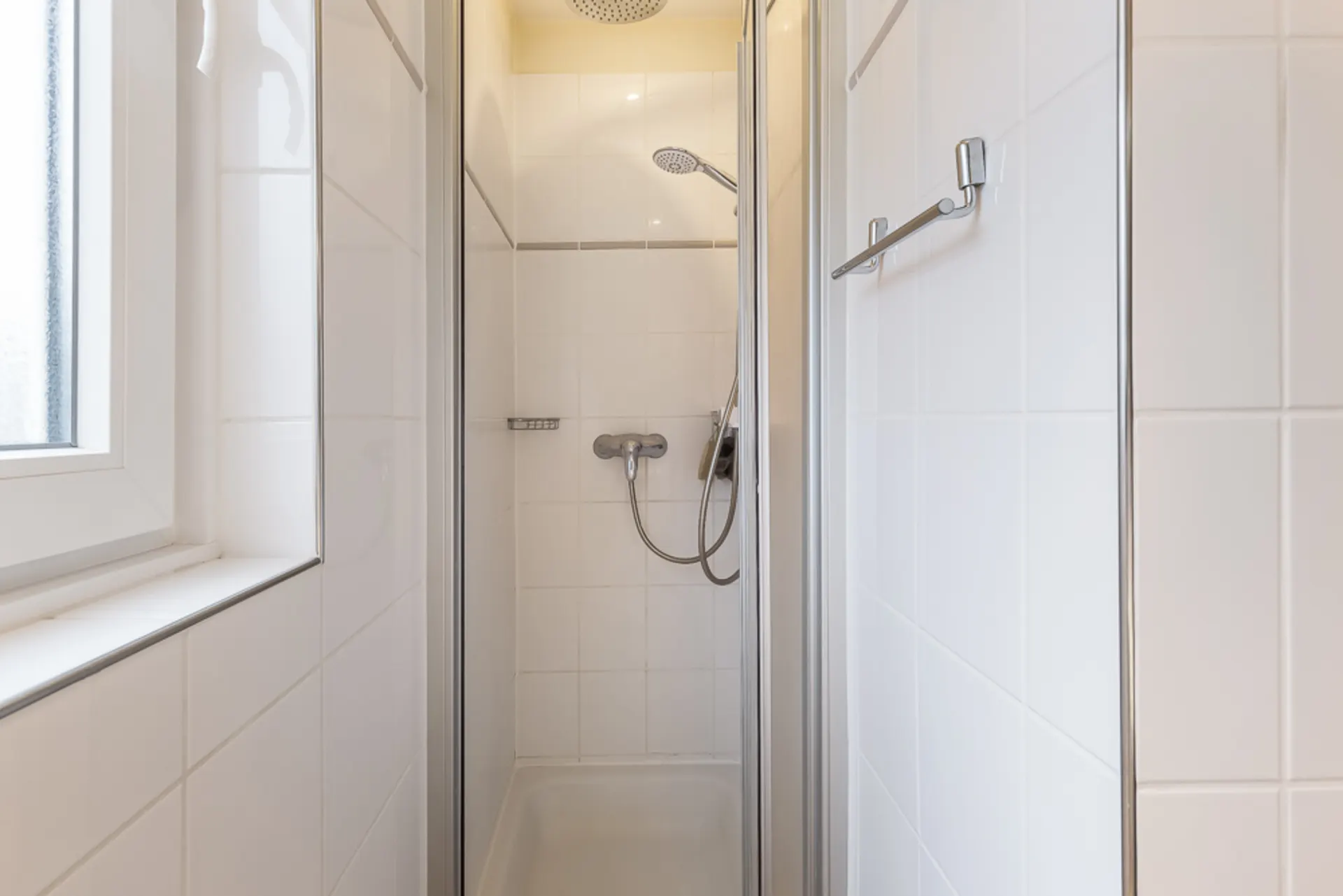 Schmale, geflieste Dusche mit Glastüren und Handbrause, neben einem Fenster.