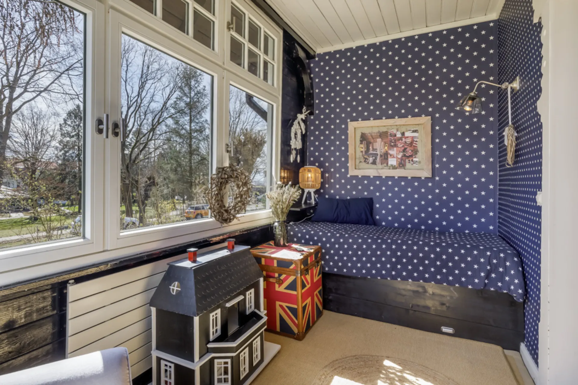 Gemütliche Sitzecke mit Sternentapete, großem Fenster, Puppenhaus und Truhe im Zimmer.