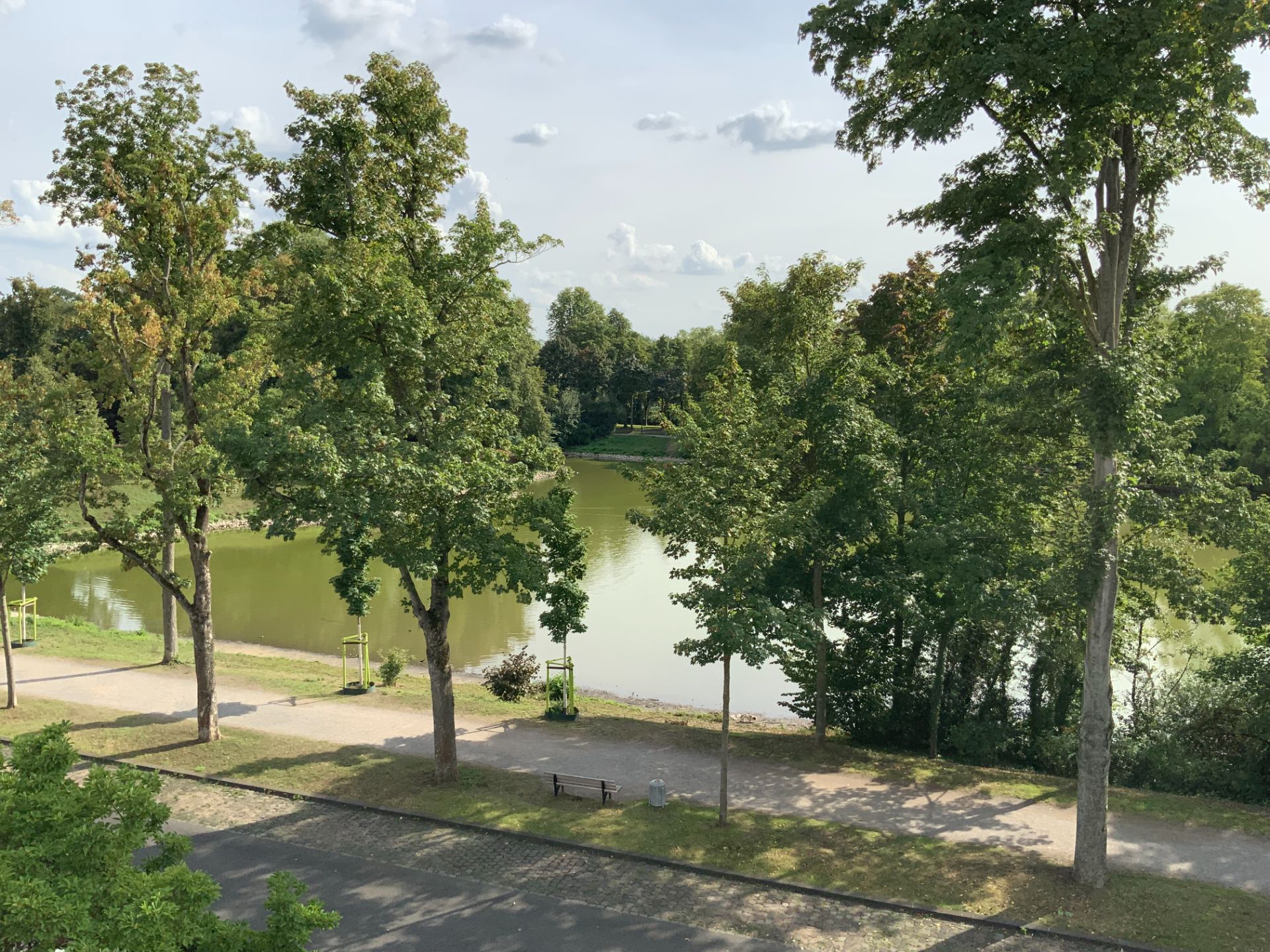 Blick auf einen grünen Park mit Bäumen, Wegen und einem ruhigen See im Hintergrund.