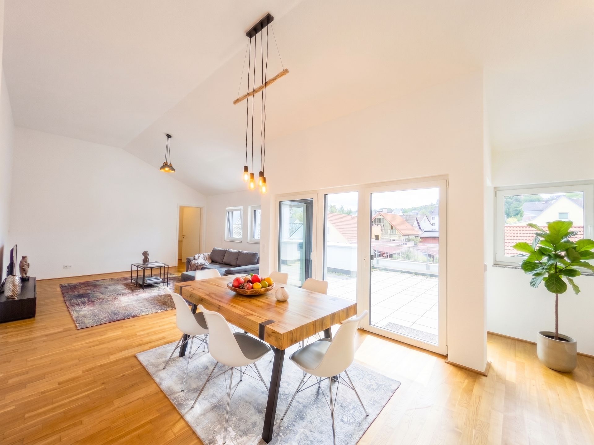 Helles Wohnzimmer mit Esstisch, vier Stühlen, Sofa, großen Fenstern und Pflanze.