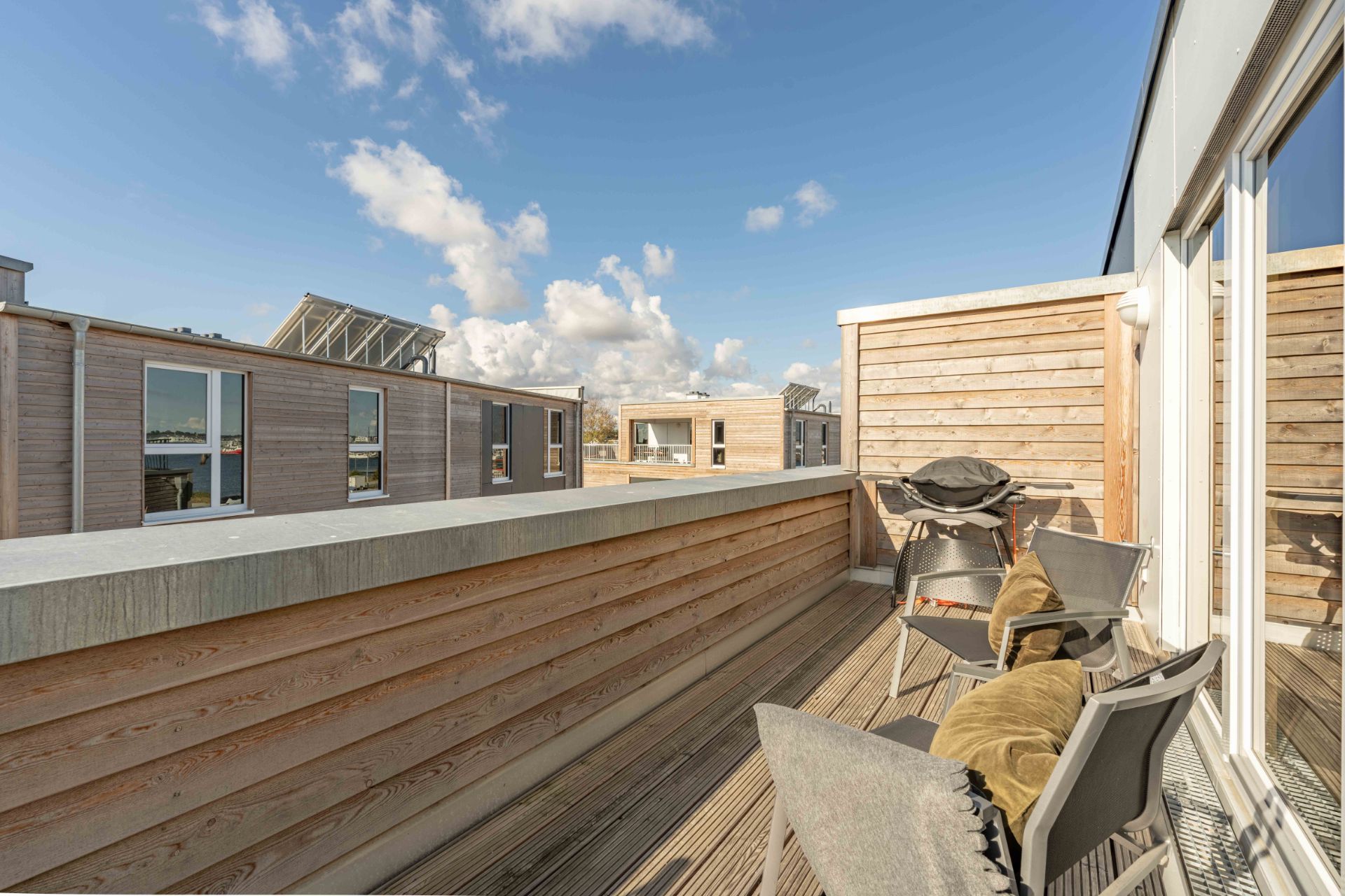 Holzterrasse mit Sitzgelegenheiten und Grill, Blick auf umliegende moderne Gebäude.