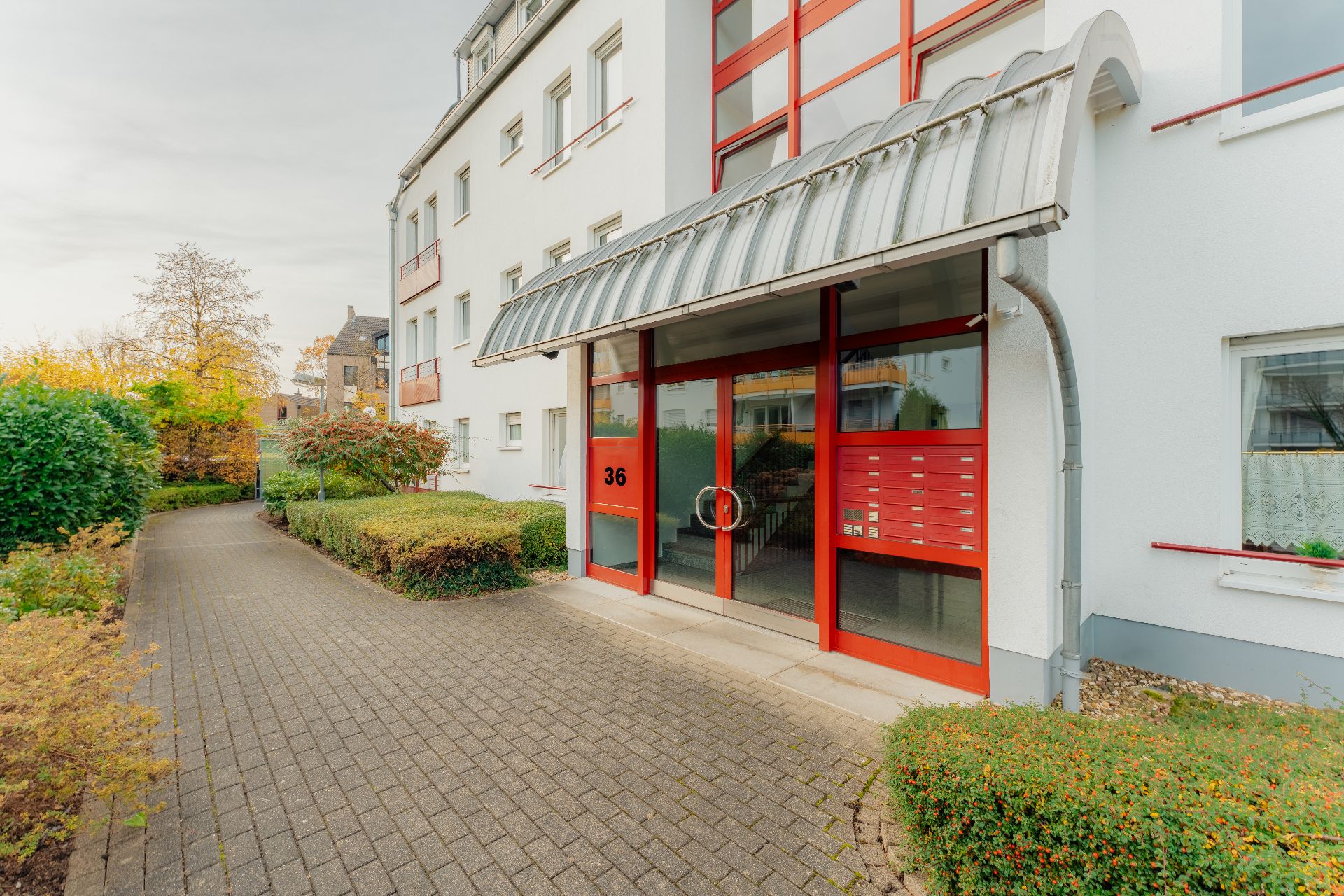 Eingang eines modernen Wohnhauses mit roten Türen und Briefkästen, umgeben von Gehweg und Grünanlagen.