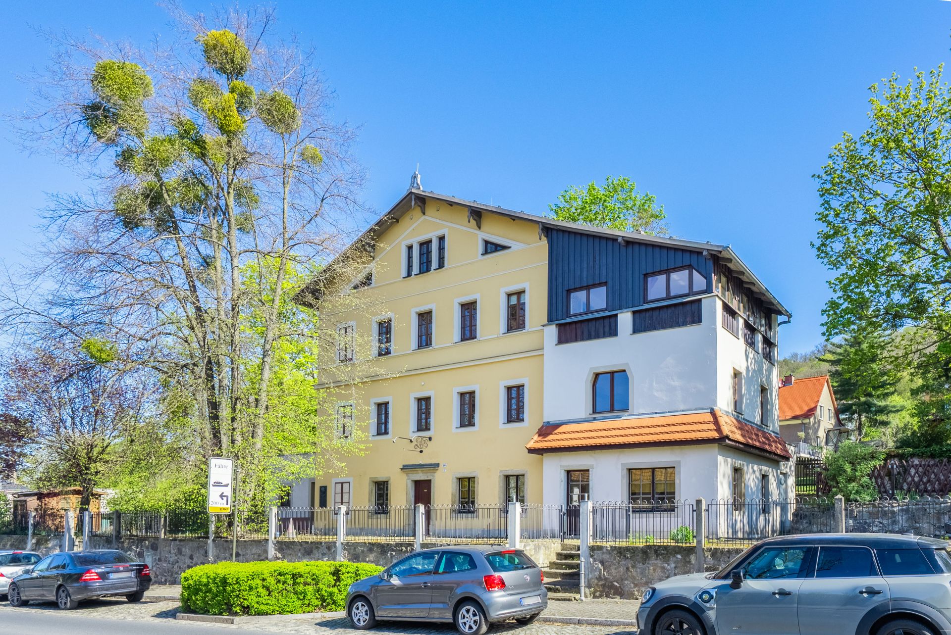 Mehrstöckiges Wohnhaus mit gelber Fassade, Autos parken davor, Bäume und blauer Himmel.