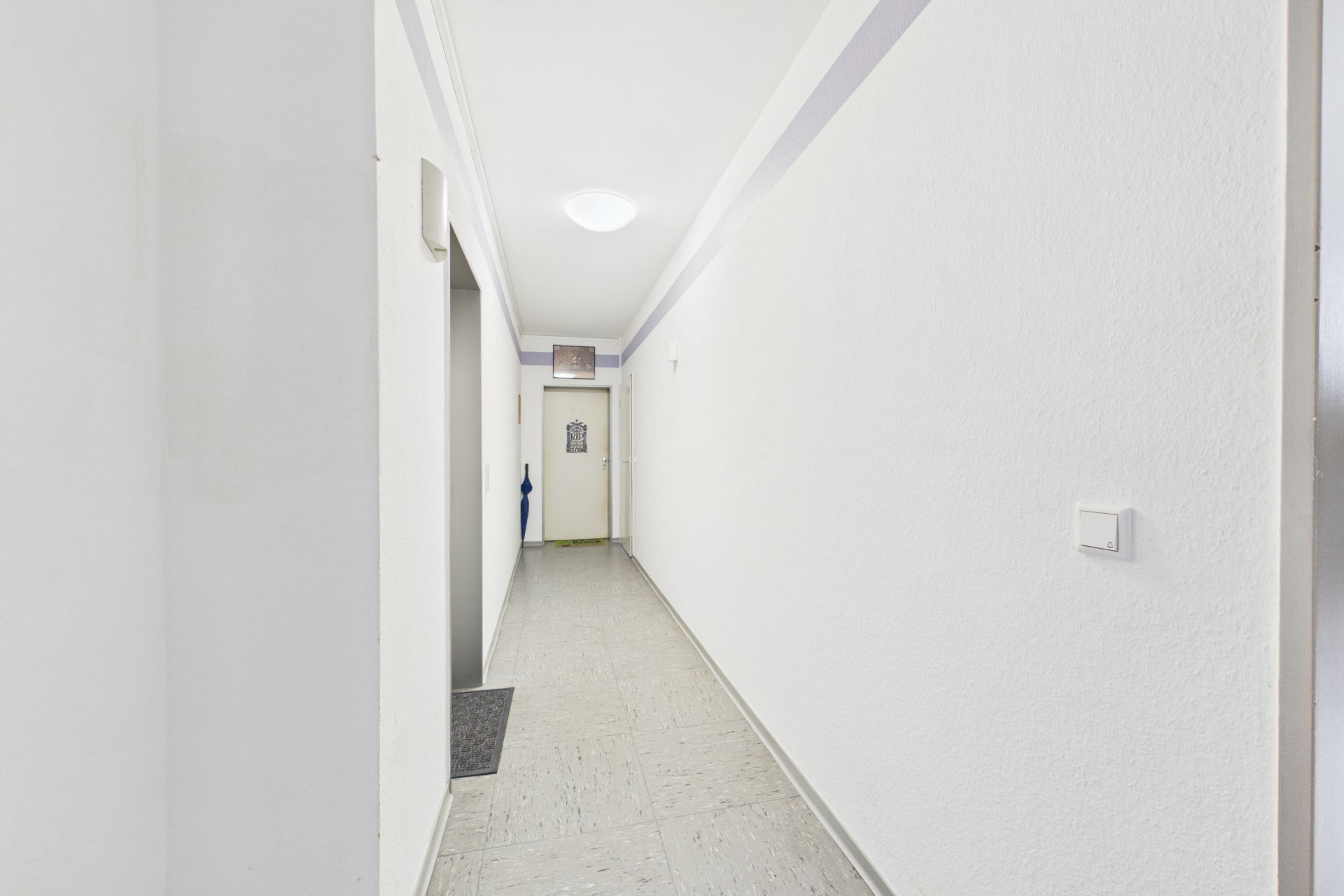 Heller Flur mit weiß gestrichenen Wänden, grauem Boden und einer Tür am Ende.