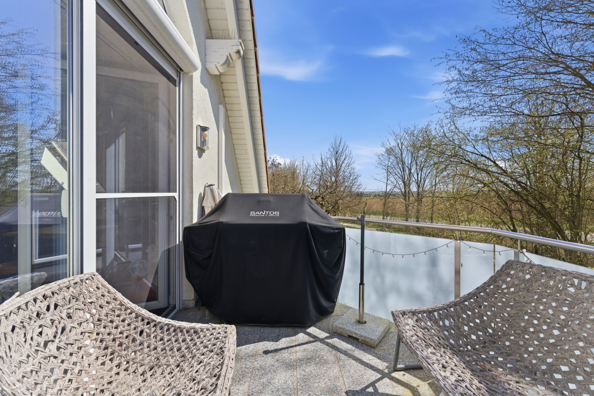 Balkon mit zwei Stühlen und einem Grill mit Abdeckung, Blick auf Bäume im Hintergrund.
