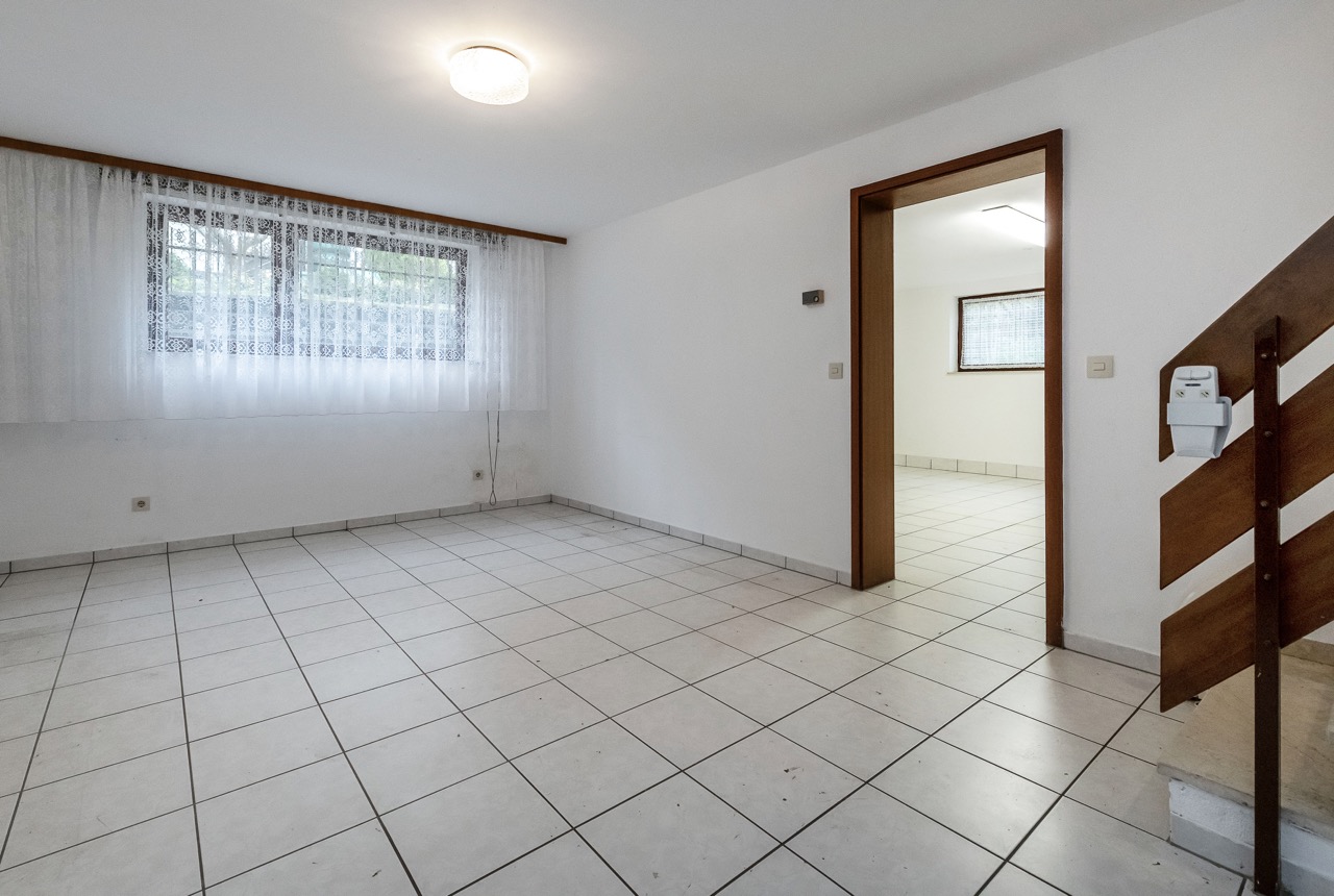 Heller, leerer Raum mit Fliesenboden, Fenster mit Gardine und angrenzendem Zimmer; Treppe rechts.