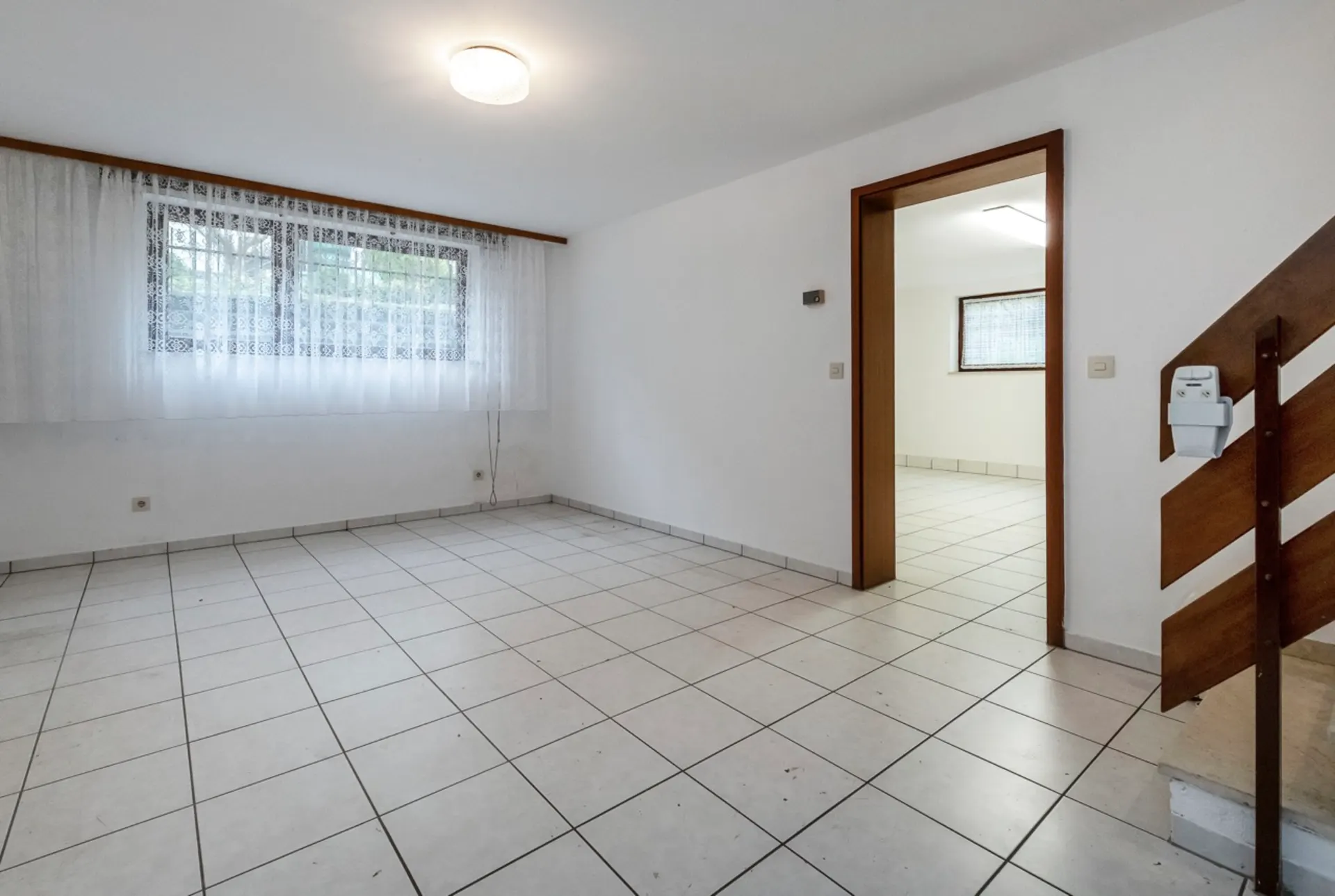 Heller, leerer Raum mit Fliesenboden, Fenster mit Gardine und angrenzendem Zimmer; Treppe rechts.