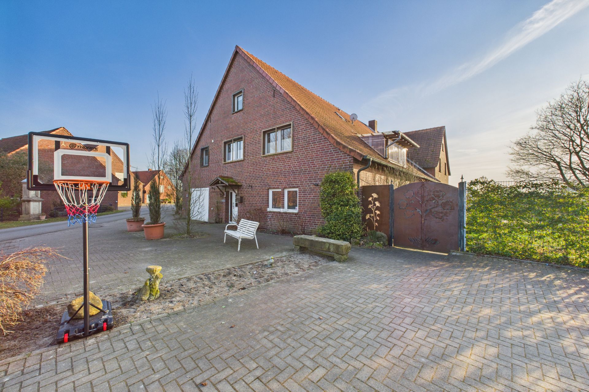 Backsteinwohnhaus mit spitzem Dach, Hof mit Basketballkorb und Bank, einige Pflanzen am Rand.