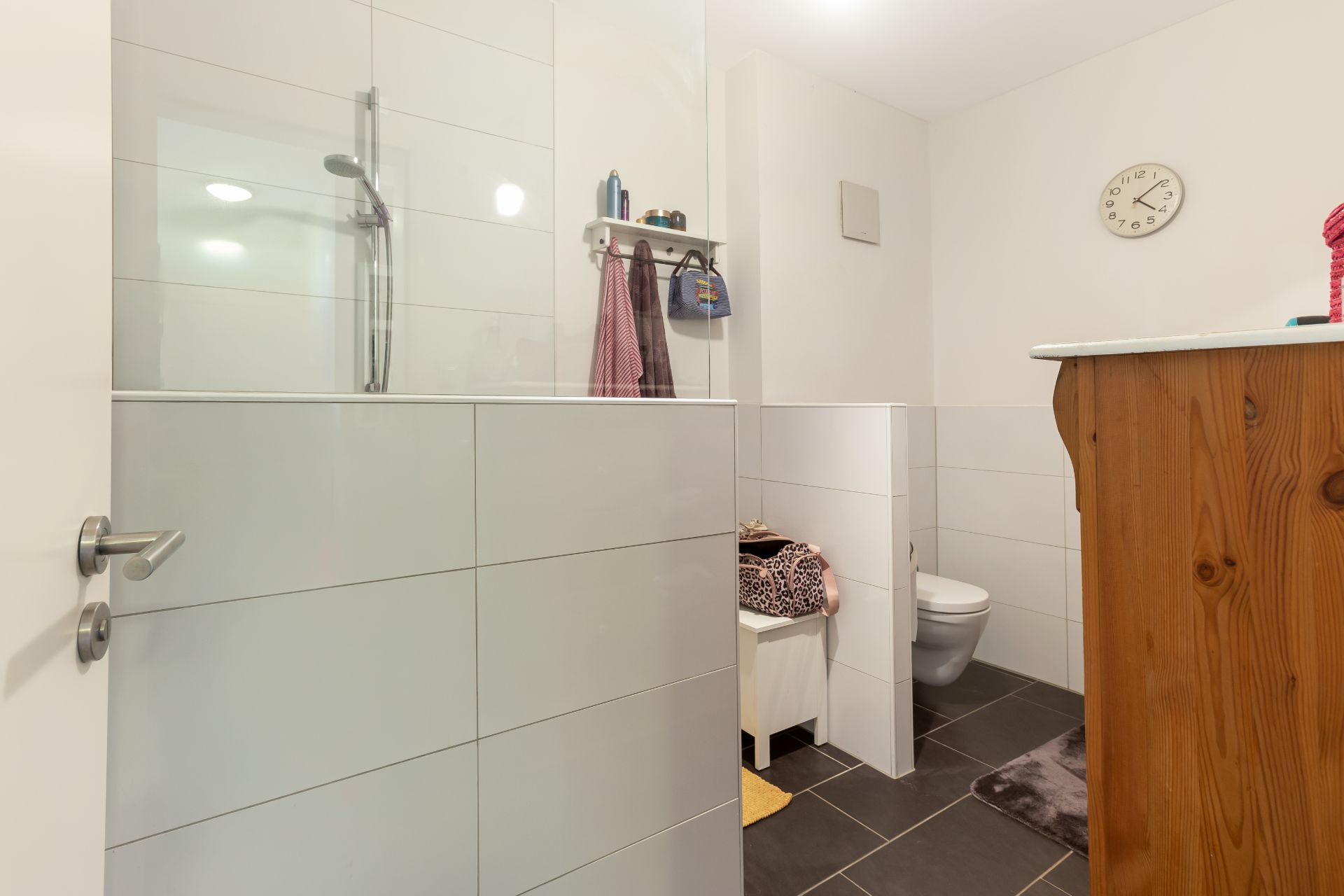 Helles Badezimmer mit offener Dusche, WC, Uhr an der Wand und einem Holzschrank.