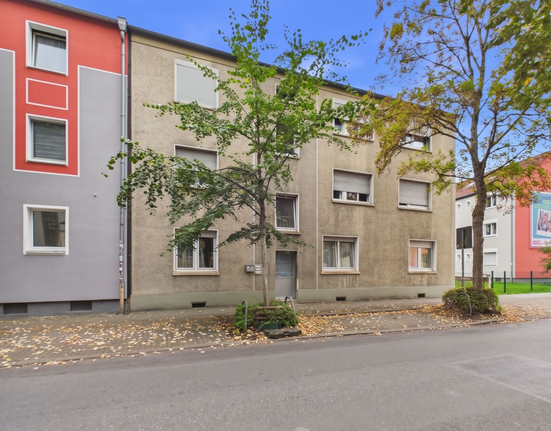 Mehrfamilienhaus mit grauer Fassade, mehreren Fenstern und Bäumen an der Straße.