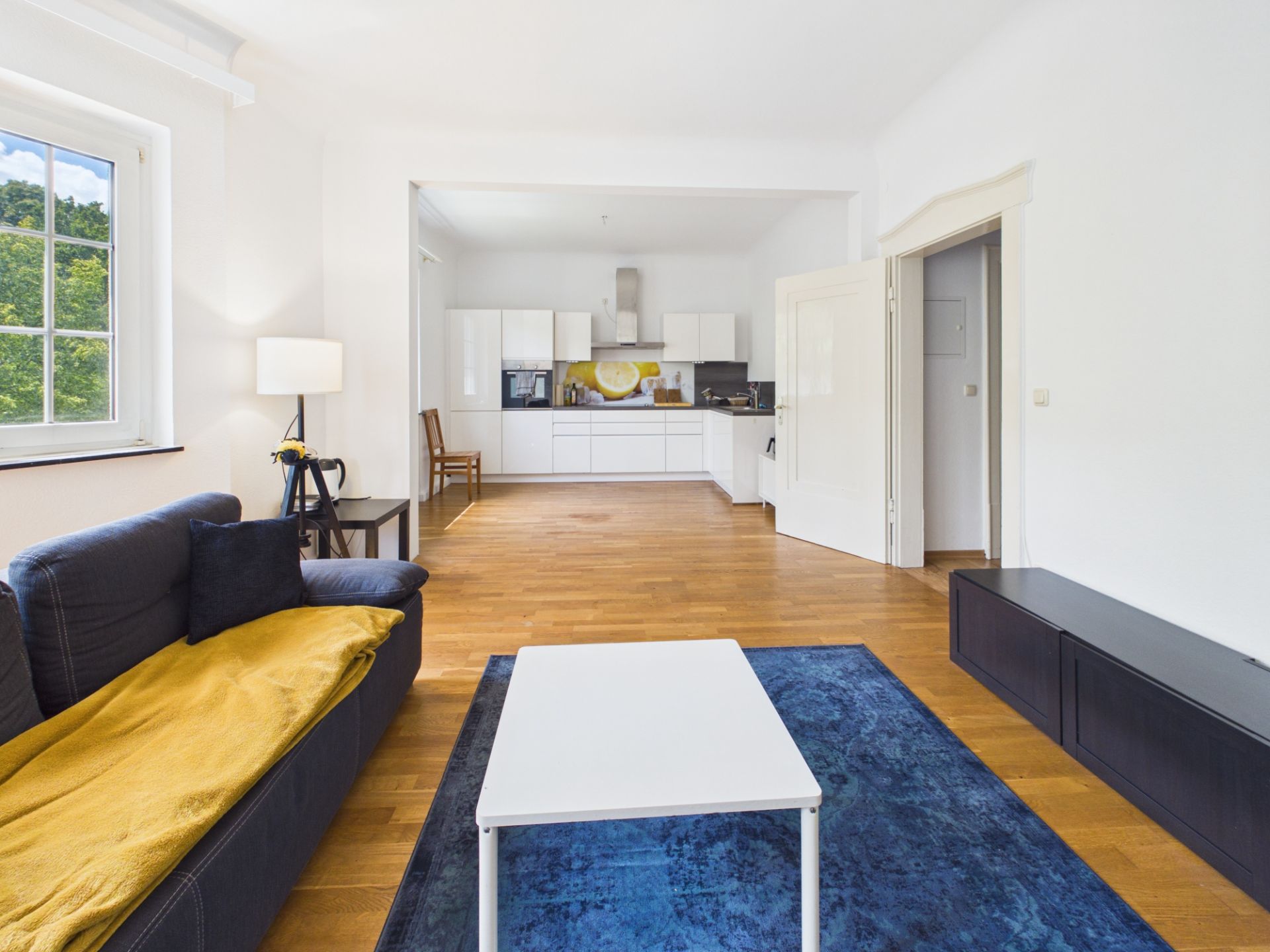 Helles Wohnzimmer mit Sofa, Couchtisch, Teppich und offener weißer Einbauküche im Hintergrund.