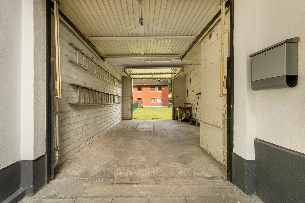 Leere Garage mit offenem Tor, Werkzeugen an der Wand und Blick auf ein rotes Wohnhaus im Hintergrund.