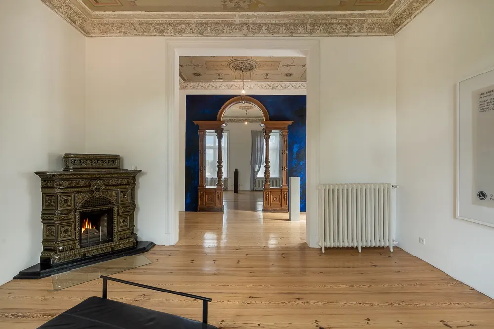 Wohnzimmer mit Kaminofen, Holzfußboden und Durchgang zu weiterem Raum mit verziertem Deckenmotiv.