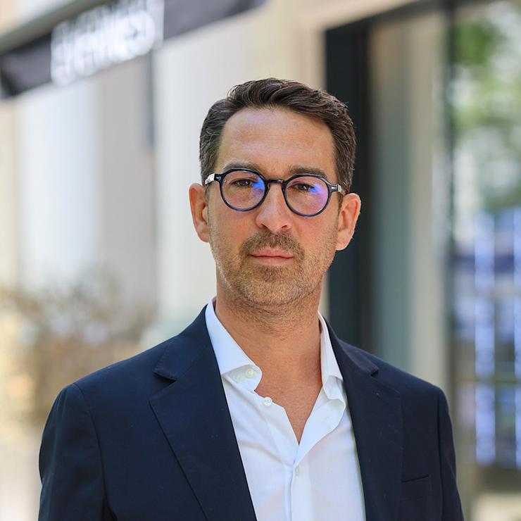 Ein Mann mit Brille und dunkelblauem Sakko steht draußen vor einem Gebäude.