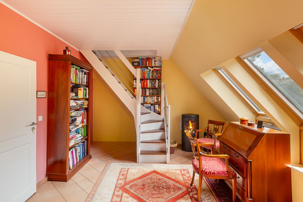 Heller Raum mit Bücherregalen, Treppe, Kaminofen und Schreibtisch unter Dachfenstern.