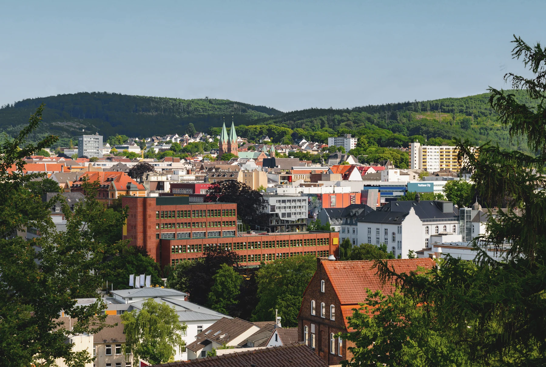 Straßenszene mit Wohnhäusern, Bäumen und einer Kirche mit zwei Türmen im Hintergrund.