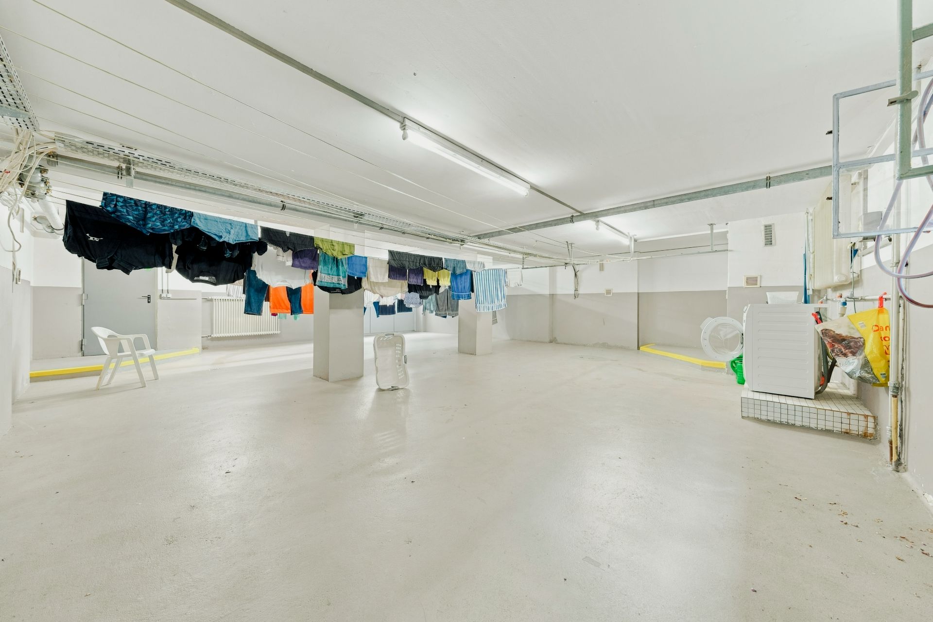 Heller Kellerraum mit Wäscheleinen und aufgehängter Kleidung, zwei weißen Stühlen und Haushaltsgegenständen.
