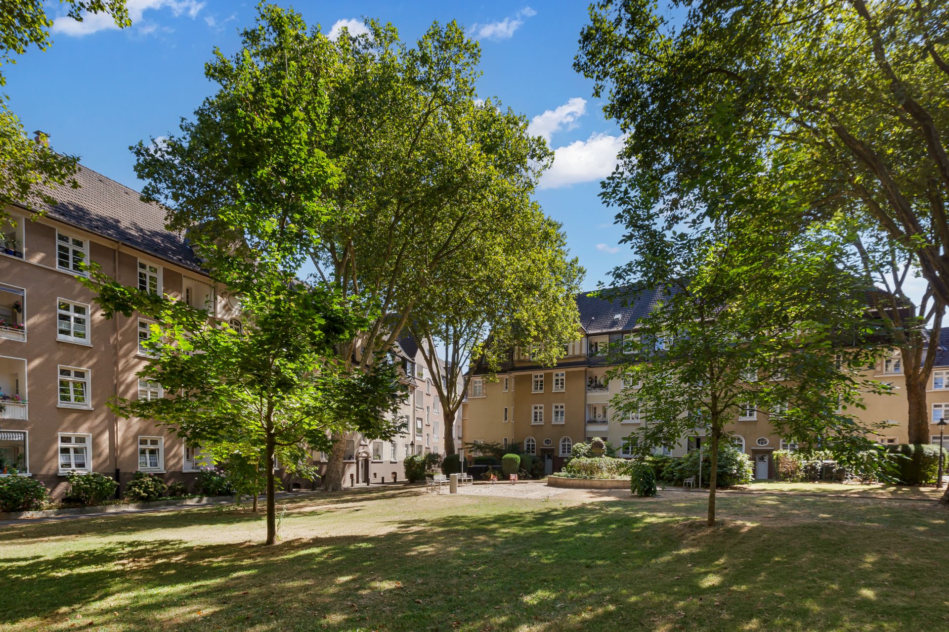 Wohngebäude mit mehreren Stockwerken, umgeben von grünem Hof und großen Bäumen an einem sonnigen Tag.