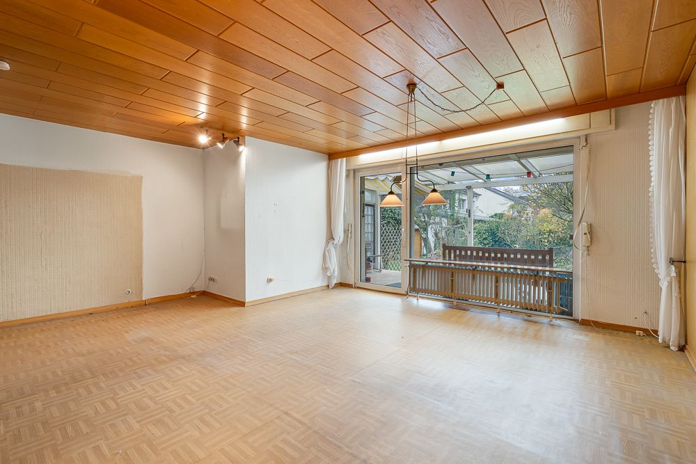 Leeres Wohnzimmer mit Holzdecke, Parkettboden und großer Fensterfront zum Garten.