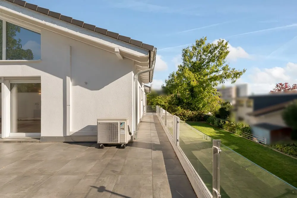 Große Terrasse mit Glasgeländer neben einem weißen Wohnhaus, Blick auf Garten und Bäume.