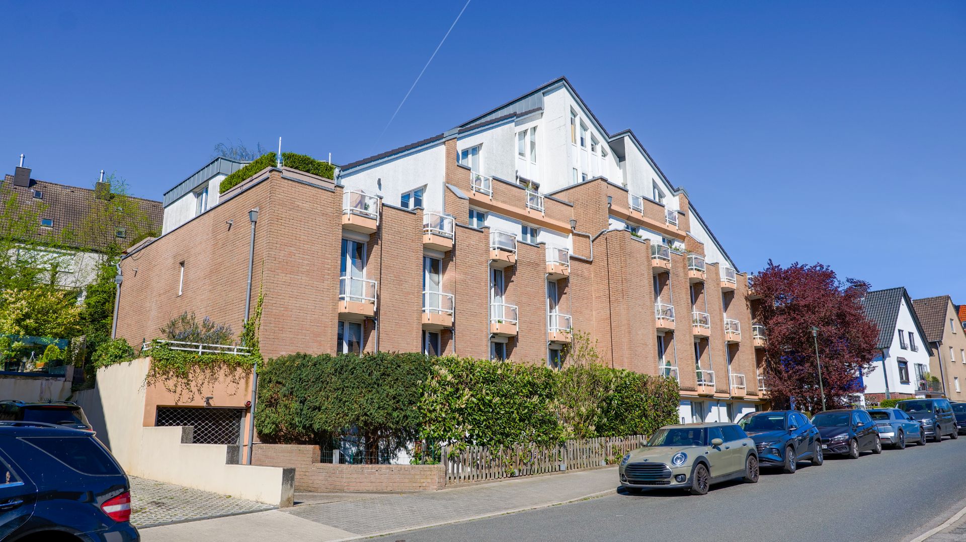 Mehrstöckiges Wohnhaus aus Backstein mit Balkonen, direkt an einer Straße mit geparkten Autos.